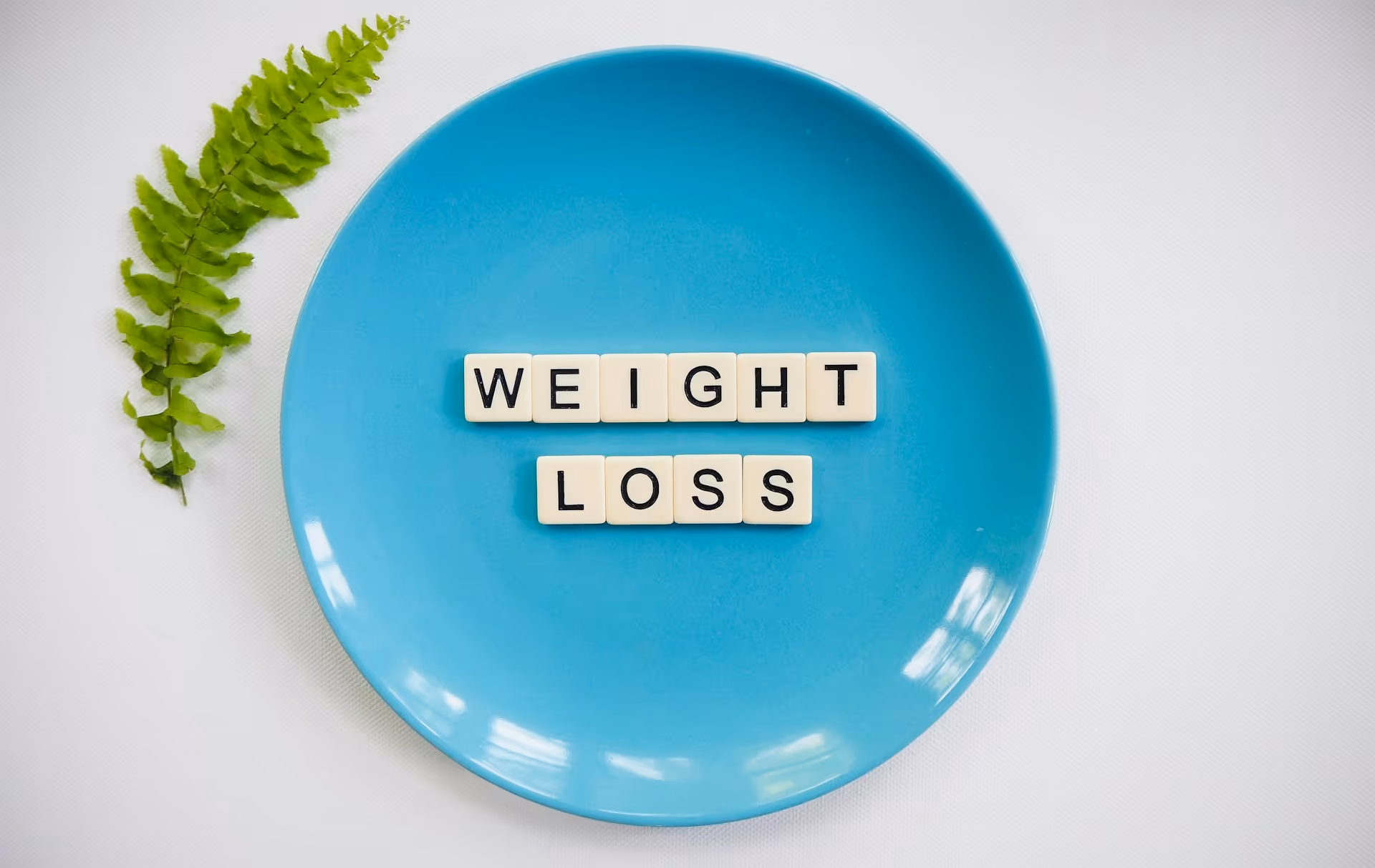 Blue plate with letter tiles spelling “WEIGHT LOSS” and a green fern leaf on a white background.