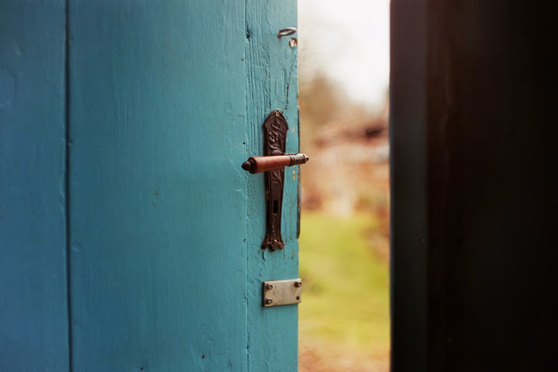A partially open turquoise door with a vintage metal handle, leading to a blurred garden background